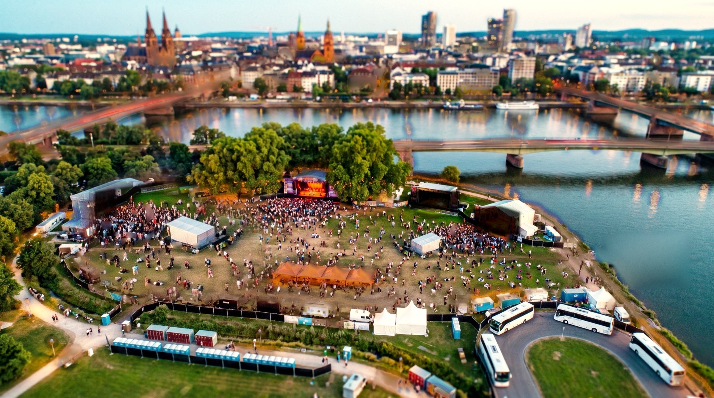 The same park in the afternoon sun: the stage under the trees lit and full, the Main Stage lawn empty between sets
